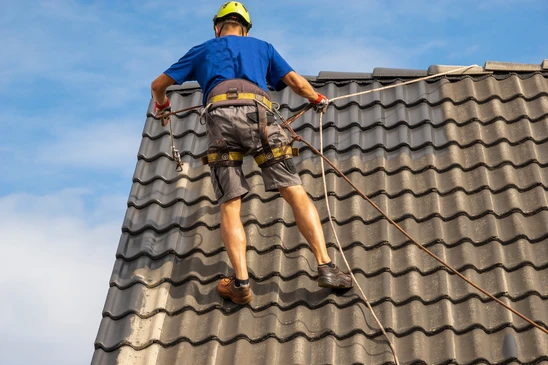 Ouvrier en harnais de sécurité travaillant sur toiture en tuiles béton ondulées, utilisant système d'ancrage avec cordes